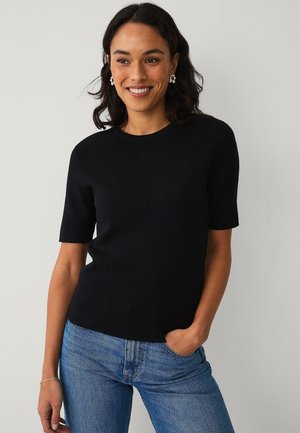 Vrouw met donker haar die zilveren oorbellen draagt, een zwart topje met korte mouwen en een blauwe spijkerbroek, glimlachend met één hand in de zak tegen een grijze achtergrond.