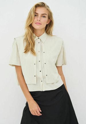 Hellbeige Kurzarmbluse mit spitzem Kragen, Druckknopfverschluss und geschichtetem Design; kombiniert mit einem schwarzen Rock.