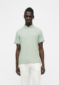 Homme portant un polo à manches courtes côtelé vert clair avec une fermeture éclair et un pantalon blanc, se tenant devant un fond blanc.