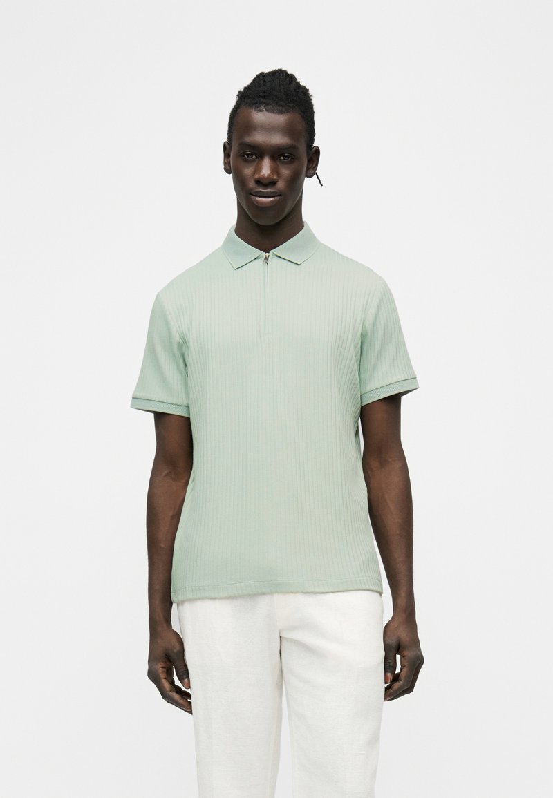 Homme portant un polo à manches courtes côtelé vert clair avec une fermeture éclair et un pantalon blanc, se tenant devant un fond blanc.