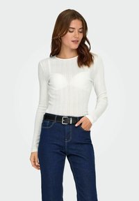 Vit ribbad långärmad topp med texturerade vertikala linjer, ihop med mörkblå denimjeans och ett svart bälte med en silverfärgad spänne.