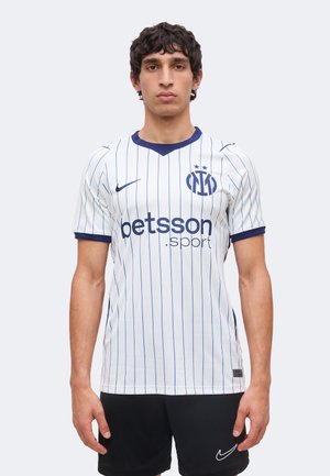 Uomo con maglia da calcio bianca e blu a righe sottili con loghi "betsson.sport" e Nike, in piedi con le braccia rilassate lungo i fianchi, rivolto in avanti.