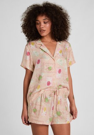 Frau mit lockigem Haar, die ein pastellrosa Kurzarmhemd mit Knöpfen und passende Shorts mit bunten Fruchtmustern trägt.