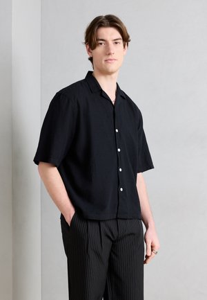 Jeune homme aux cheveux bruns mi-longs portant une chemise noire à manches courtes boutonnée et un pantalon noir à fines rayures, debout les mains dans les poches.