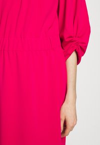 Bras droit et main d'une personne portant une robe rose vif à manches longues avec un détail froncé au poignet et à la taille.