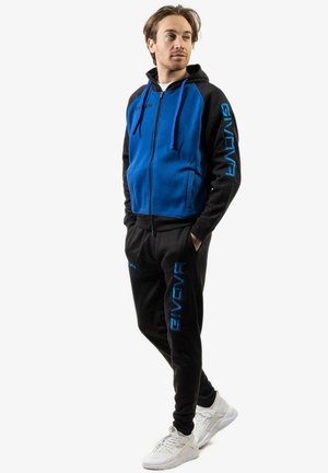 Modello maschile in felpa con zip blu e nera e pantaloni sportivi neri abbinati con loghi GIVOVA blu, in piedi con le mani in tasca, mentre guarda a destra.