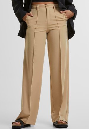 Beige high-waisted wijde broek met plooien, voorzien van zijzakken en een gladde textuur. Gecombineerd met zwarte platform sandaal.