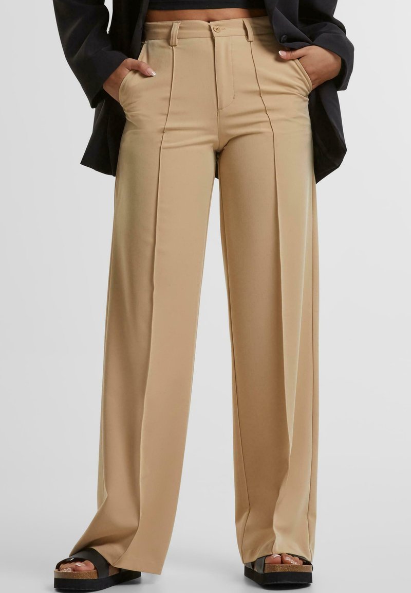 Beige high-waisted wijde broek met plooien, voorzien van zijzakken en een gladde textuur. Gecombineerd met zwarte platform sandaal.