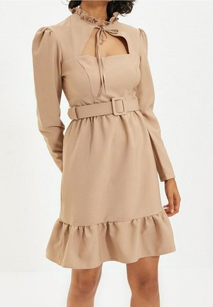 Beiges langärmeliges Kleid mit Rüschenkragen, quadratischem Ausschnitt, tailliert mit Gürtel und gerüschtem Saum, getragen von einer stehenden Person.