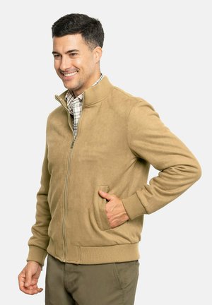 Chaqueta bomber color tan de material suave y texturizado, con cierre de cremallera, puños acanalados y un bolsillo lateral. Usada sobre una camisa de cuadros.