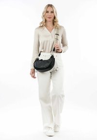 Sort læderhandbag med en rund form, der har en tekstureret overflade og en mønstret justerbar rem. Model iført cremefarvet knap-op skjorte og hvide jeans.
