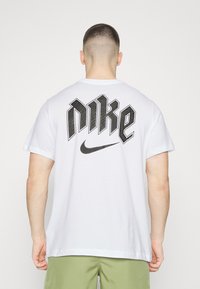 Camiseta de algodón blanca con un gran logo negro de "Nike" en diseño tridimensional en la parte posterior. Mangas cortas y cuello redondo.
