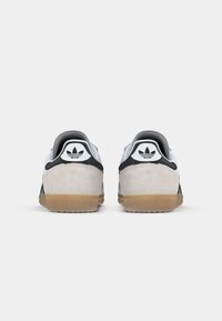 Par de ténis Adidas brancos e pretos com solas de borracha mostrados de trás, exibindo o logo trevo da Adidas no calcanhar.