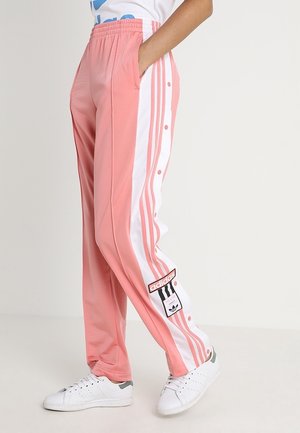 Osoba nosząca różowe dresy Adidas z białymi bocznymi paskami i zatrzaskami, w połączeniu z białymi tenisówkami.