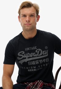Camiseta de algodón negra con un gran gráfico gris que incluye el texto "SuperDry" y caracteres japoneses. Diseño de manga corta y cuello redondo.