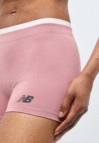 Κοντινό πλάνο ατόμου που φοράει ροζ αθλητικές σορτς New Balance με το χέρι να ακουμπά στο μηρό, σε ανοιχτόχρωμο φόντο.