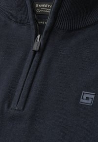 Gros plan d'un pull en maille bleu marine foncé avec un col demi-fermeture éclair et un petit logo brodé sur la poitrine.