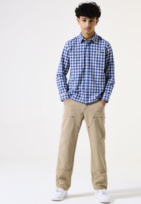 Camisa de manga larga a cuadros en azul y blanco con dos bolsillos en el pecho, combinada con pantalones beige claros que tienen dobles bolsillos frontales y zapatillas blancas.