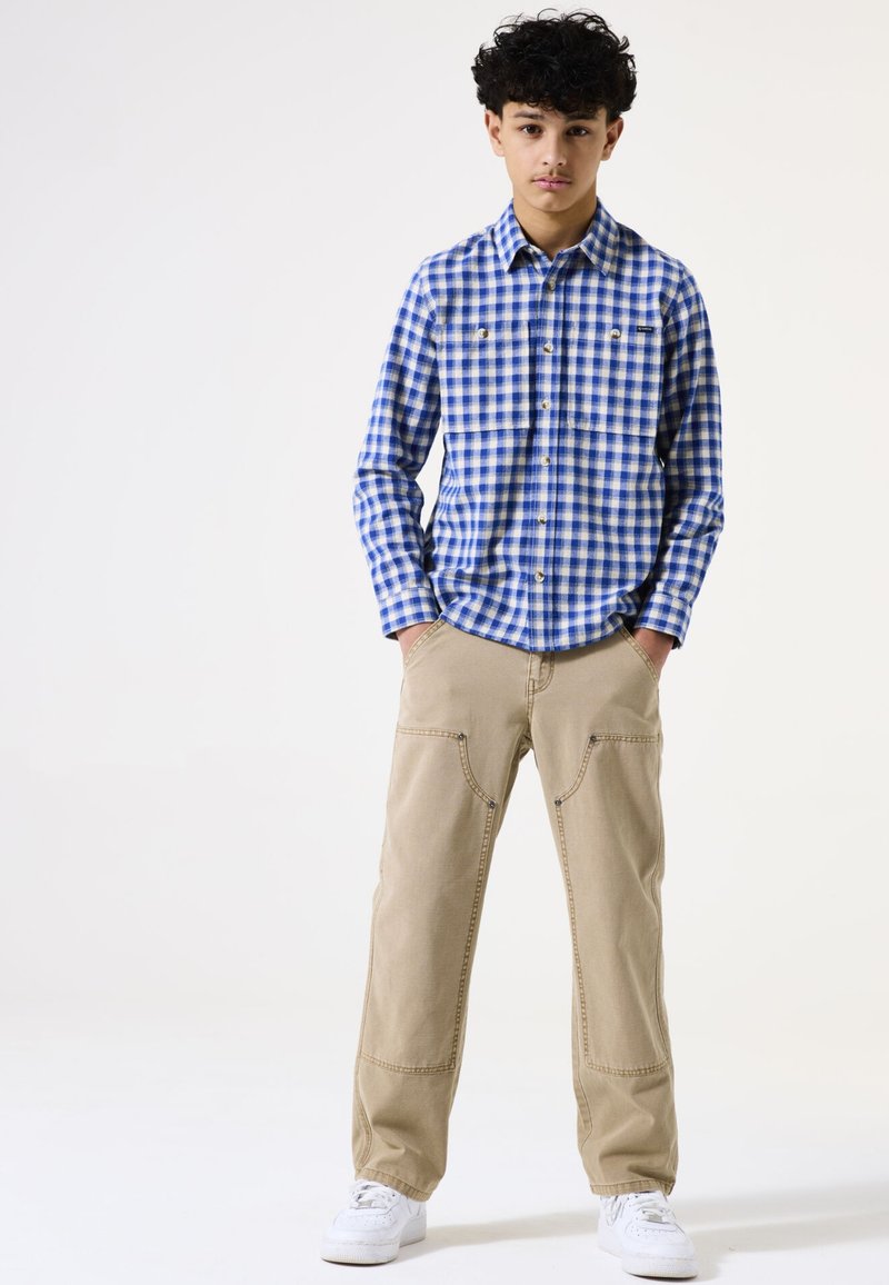 Camisa de manga larga a cuadros en azul y blanco con dos bolsillos en el pecho, combinada con pantalones beige claros que tienen dobles bolsillos frontales y zapatillas blancas.