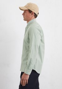 Langärmliges Hemd mit grün-weißen Streifen, mit geknöpften Manschetten und Kragen. Trägt eine beigefarbene Mütze. Einfaches und figurbetontes Design.