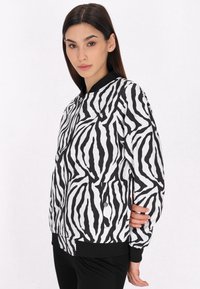 Bunda bomber s zebrovým vzorem v černobílé barvě, s dlouhými rukávy, příjemným ribbed límcem a manžetami, a předními kapsami se zapínáním na cvočky.