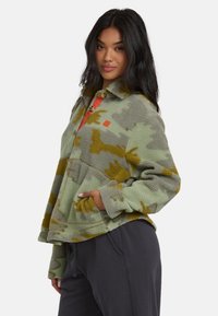 Donna con lunghi capelli scuri, che indossa una giacca in pile a motivo verde e marrone e pantaloni grigio scuro, in piedi con una mano in tasca.