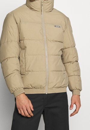 Homme portant une doudoune beige avec col montant et poignets élastiques, associée à un pantalon noir, sur un fond uni clair.