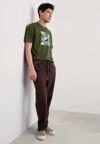 Žalias pieštas T-shirt su geometriniu dizainu šviesiai mėlyna spalva; derinamas su tamsiai rudomis kelnėmis ir smėlio spalvos sportbačiais. Kasdienis siluetas ir stilius.