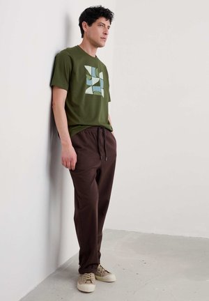 Groene graphic T-shirt met een geometrisch ontwerp in lichtblauw; gecombineerd met donkere bruine broek en beige sneakers. Casual fit en stijl.