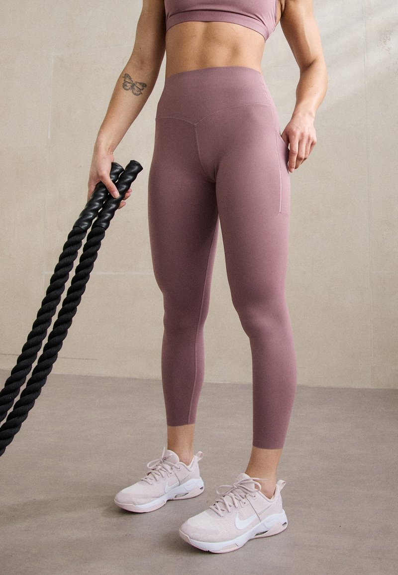 Leggings a vita alta di un rosa polveroso con una texture liscia e una tasca laterale, abbinati a scarpe da ginnastica rosa chiaro. Corda da esercizio nera tenuta in mano.