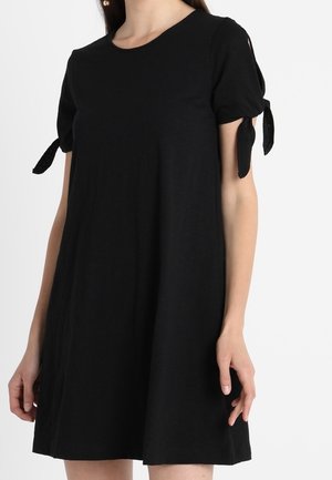 Robe noire à manches courtes avec encolure ronde et nœuds noués sur les manches, portée par une personne avec les bras détendus le long du corps.