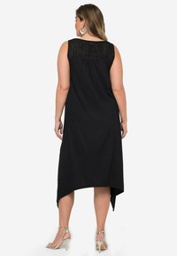 Schwarzes ärmelloses Kleid mit einem Spitzenmuster auf der Rückseite, mit einem hoch-niedrig Saum und einer fließenden Silhouette, kombiniert mit Riemchenabsätzen.