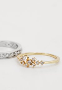 Anello d'oro con numerosi piccoli pietre chiare in design a grappolo, accanto a un anello d'argento con una fila di pietre chiare incastonate.