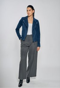 Marineblauwe suède motorjas met een asymmetrische ritssluiting, gecombineerd met lichtgrijze wijde broekspijpen en zwarte puntige enkellaarsjes.