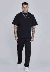 Czarny T-shirt z krótkim rękawem z małym białym logo, zestawiony z czarnymi spodniami i białymi trampkami, o luźnym kroju i gładkiej fakturze.