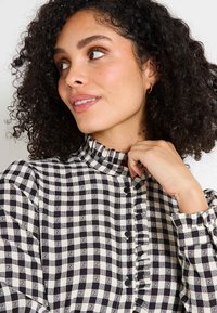 Mujer con cabello rizado que lleva una blusa a cuadros en blanco y negro con botones y cuello con volantes, mirando hacia un lado, con la mano cerca del cuello.