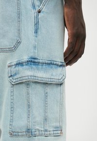 Világoskék farmer overall, nagy, varrt zsebbel a combján, kontrasztos narancssárga varrással és kopott textúrával.