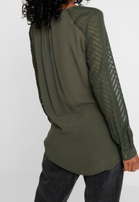 Donna che indossa una blusa verde oliva con maniche lunghe trasparenti ricamate e dettaglio a chiave nella parte posteriore, abbinata a pantaloni grigio scuro.