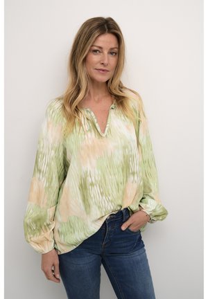 Blouse à motifs vert et pêche avec un col en V, des manches ballon longues et une coupe ample. Portée avec un jean bleu foncé.