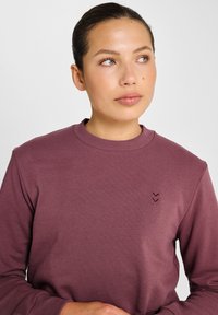 Paarse trui met een ronde hals en geribbelde boorden. Voorzien van een klein chevron-logo op de borst. Gladde, zachte stoftextuur.