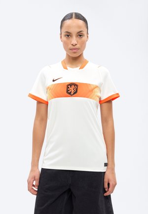 Νεαρή γυναίκα που φοράει λευκή και πορτοκαλί φανέλα ποδοσφαίρου Nike με έμβλημα λιονταριού, στέκεται μπροστά σε ένα απλό φωτεινό φόντο.