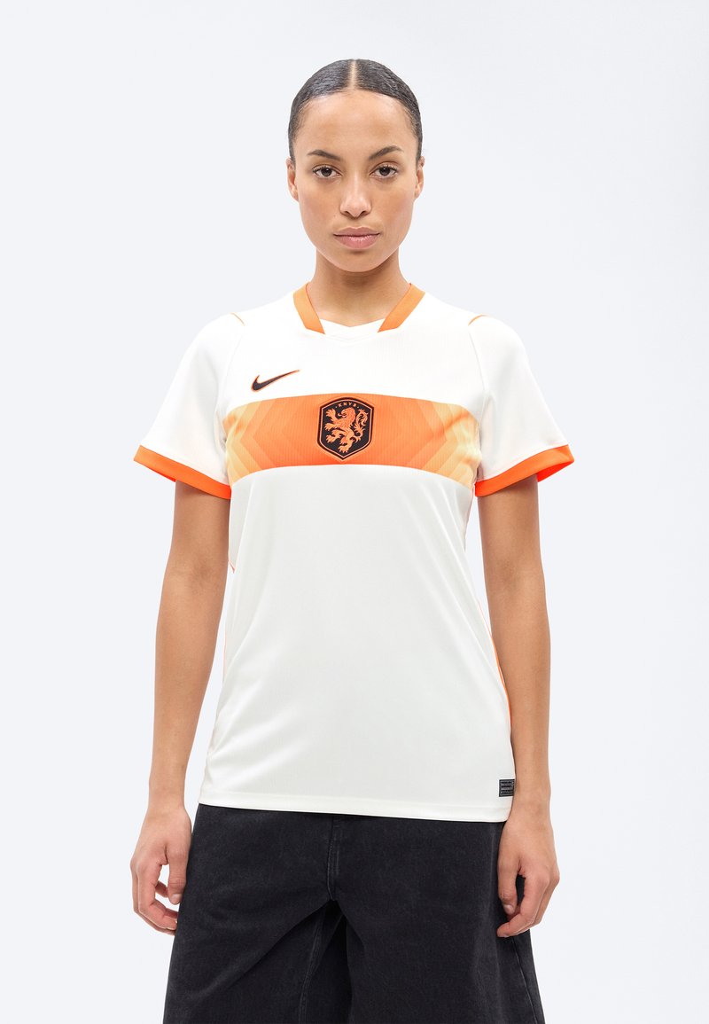 Νεαρή γυναίκα που φοράει λευκή και πορτοκαλί φανέλα ποδοσφαίρου Nike με έμβλημα λιονταριού, στέκεται μπροστά σε ένα απλό φωτεινό φόντο.