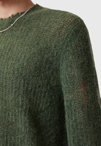 Zielony sweter dziergany z teksturowanym wzorem, z wykończeniem brzegów dekoltu na surowo oraz małym zniszczeniem na jednym z rękawów. Miękki, puszysty materiał.