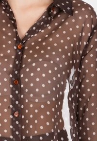 Camicia trasparente marrone con bottoni bianchi a pois, dotata di colletto classico e accenti di bottoni arancioni. Texture liscia, tessuto leggero.