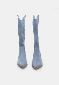 Blå mockaboots med spetsiga tår, dekorativa utskärningar, dragkedjor på sidorna och en strukturerad yta.