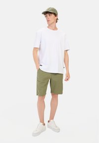 Hvid kortærmet t-shirt, olivengrønne shorts og hvide sneakers; mand står med hånden i lommen, iført en grøn kasket.