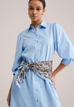 Blå bomuldsskjortekjole med puffede ærmer, knaplukning og et brunt og hvidt slangemønstret tørklæde bundet om taljen.