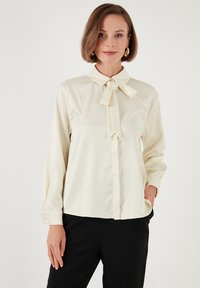Blusa blanca cremosa de tela suave, con cuello y detalle de lazo, mangas largas y puños abotonados.