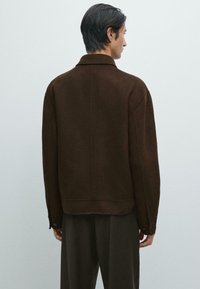 Brun uldblandingsjakke med en afslappet pasform, subtil tekstur, foldet krave og knaplukninger på manchetterne, set bagfra mod en lys baggrund.