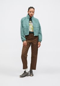 Jasnoturkusowa teksturowana kurtka z wysokim kołnierzem, zestawiona z koszulką z nadrukiem, brązowymi spodniami oraz szarymi tenisówkami z jasnymi podeszwami.